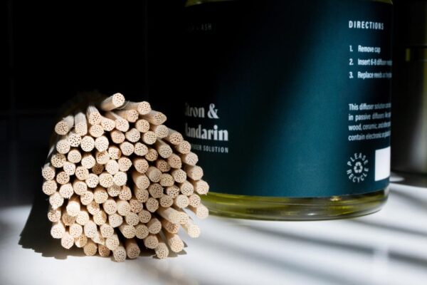 A close-up shot of a bundle of natural rattan diffuser reeds next to a bottle of Windsor + Ash Citron & Mandarin diffuser solution, clearly showing the usage directions on the label.