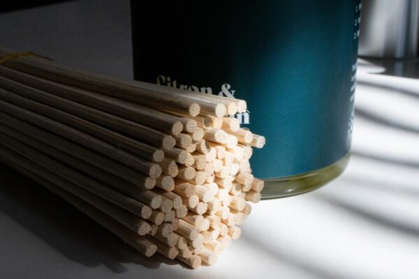 A close-up view of a bundle of natural rattan diffuser reeds beside a dark bottle of Windsor + Ash Citron & Mandarin diffuser solution, featuring a subtle shadow pattern.