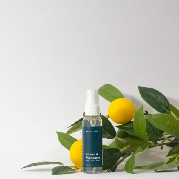 A small, clear bottle of Windsor + Ash Citron & Mandarin Room & Linen Spray with a white sprayer, positioned in front of green leafy branches and two vibrant yellow lemons, against a clean white background.